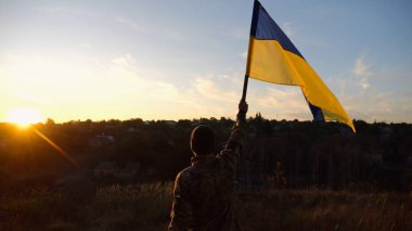 Erkek asker, güzel günbatımının arka planında Ukrayna bayrağıyla ayakta duruyor. Rus saldırısına karşı kazanılan zaferin şerefine mavi-sarı bayrak taşıyan Ukraynalı erkek asker..