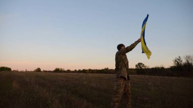 Genç erkek ordu üniformalı Ukrayna bayrağıyla alacakaranlıkta çayırda koşuyor. Ukrayna ordusunun askeri Rus saldırısına karşı zafer şerefine mavi-sarı bayrak sallıyor. Savaşın sonu. Yavaş çekim