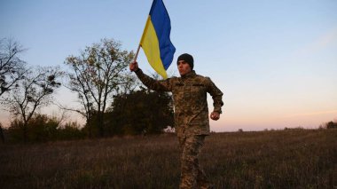 Alacakaranlıkta mavi-sarı bayrak taşıyan Ukrayna ordusunun askeri. Çayırda Ukrayna bayrağıyla koşan üniformalı genç bir erkek asker. Rus saldırganlığına karşı zafer.