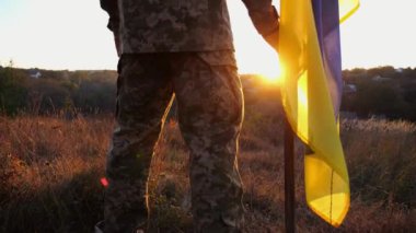 Ukraynalı asker gün batımında kırsal bölgede ulusal bayrağıyla ayakta duruyor. Rus saldırısına karşı zafer sembolü olarak Ukrayna bayrağı taşıyan askeri üniformalı bir erkek asker. İstila direnişi kavramı