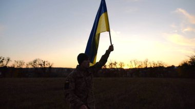 Ukrayna ordusunun askeri gün batımında mavi-sarı pankartıyla sahaya çıkıyor. Üniformalı genç erkek asker çayırda Ukrayna bayrağıyla yürüyor. Rus saldırısına karşı zafer kavramı.