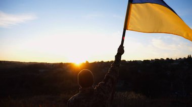 Erkek asker, güzel günbatımının arka planında Ukrayna bayrağıyla ayakta duruyor. Rus saldırısına karşı kazanılan zaferin şerefine mavi-sarı bayrak taşıyan Ukraynalı erkek asker..