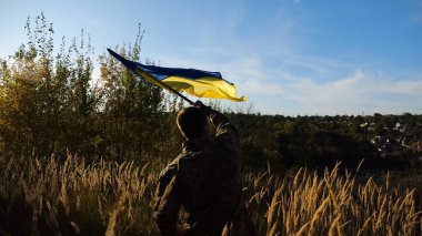 Kırsal kesimde Ukrayna bayrağı sallayan üniformalı bir erkek asker. Ukrayna ordusunun genç askeri Rus saldırısına karşı zafer sembolü olarak mavi-sarı bayrağı kaldırıyor. İstila direnişi kavramı