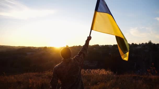 カモフラージュのユニフォームの若者は ロシアの侵略に対する勝利を記念して青い黄色の旗を持ち上げた 男性のウクライナ軍兵士がウクライナの波状旗を掲げ 背景に日没した — ストック動画