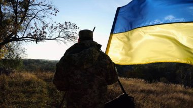 Üniformalı genç erkek asker Ukrayna bayrağıyla kırsalda yürüyor. Ukrayna ordusunun askeri açık havada mavi-sarı bayrak sallıyor. Rus saldırısına karşı zafer kavramı