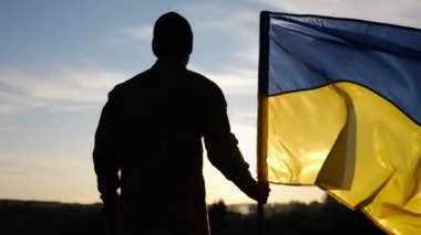 Ukraynalı asker gün batımında kırsal bölgede ulusal bayrağıyla ayakta duruyor. Rus saldırısına karşı zafer sembolü olarak Ukrayna bayrağı taşıyan askeri üniformalı bir erkek asker. İstila direnişi kavramı
