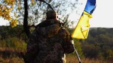 Üniformalı genç erkek asker Ukrayna bayrağıyla kırsalda yürüyor. Ukrayna ordusunun askeri açık havada mavi-sarı bayrak sallıyor. Rus saldırısına karşı zafer kavramı.