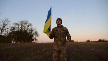 Alacakaranlıkta mavi-sarı bayrak taşıyan Ukrayna ordusunun askeri. Çayırda Ukrayna bayrağıyla koşan üniformalı genç bir erkek asker. Rus saldırganlığına karşı zafer.