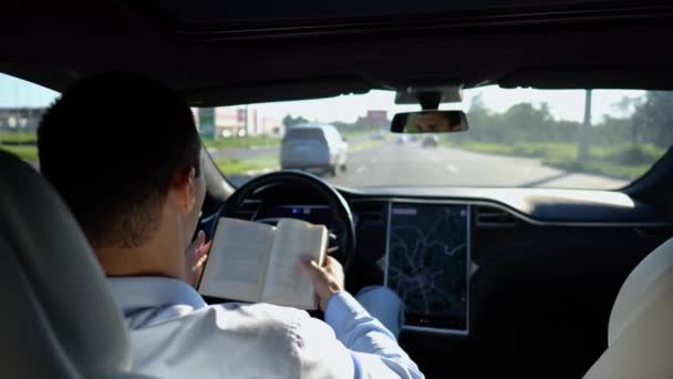 在城市道路上驾驶自动驾驶仪的电动汽车时 男性商人在看书 成功的商人在驾驶自动驾驶电动汽车的同时提高了自己的知识水平 — 图库视频影像