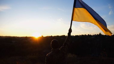 Erkek asker, güzel günbatımının arka planında Ukrayna bayrağıyla ayakta duruyor. Rus saldırısına karşı kazanılan zaferin şerefine mavi-sarı bayrak taşıyan Ukraynalı erkek asker..