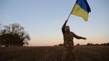 Genç erkek ordu üniformalı Ukrayna bayrağıyla alacakaranlıkta çayırda koşuyor. Ukrayna ordusunun askeri Rus saldırısına karşı zafer şerefine mavi-sarı bayrak sallıyor. Savaşın sonu. Yavaş çekim