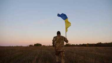 Alacakaranlıkta mavi-sarı bayrak taşıyan Ukrayna ordusunun askeri. Çayırda Ukrayna bayrağıyla koşan üniformalı genç bir erkek asker. Rus saldırganlığına karşı zafer.