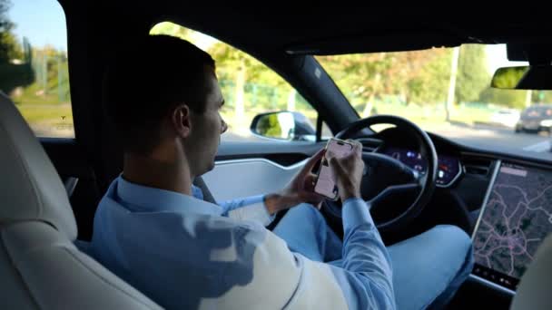 成功したビジネスマンは 自動運転の電気自動車に乗っている間 現代のスマートフォンを閲覧します 都市道路での自動操縦で電気自動車に乗っている間に携帯電話を使用する男性起業家 — ストック動画