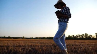 Gün batımında buğday çayırlarında dolaşırken dijital tablet kullanan dişi tarım uzmanı. Çiftçi arpa tarlasında yürürken hasatı izliyor. Güzel manzara manzarası. Tarım sektörü kavramı.
