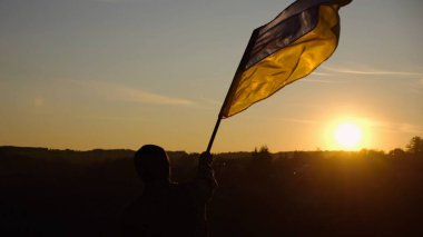 Askeri üniformalı genç adam arka planda güzel günbatımına karşı Ukrayna bayrağı sallıyor. Erkek Ukraynalı asker kırsal kesimde ulusal bayrağı kaldırdı. Rus saldırısına karşı zafer.