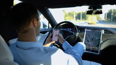 Başarılı bir iş adamı, otonom elektrikli araba kullanırken modern akıllı telefona göz atıyor. Şehir yolunda otomatik pilotla elektrikli araba sürerken telefon kullanan erkek girişimci..