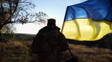 Üniformalı genç erkek asker Ukrayna bayrağıyla kırsalda yürüyor. Ukrayna ordusunun askeri açık havada mavi-sarı bayrak sallıyor. Rus saldırısına karşı zafer kavramı.