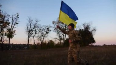 Alacakaranlıkta mavi-sarı bayrak taşıyan Ukrayna ordusunun askeri. Çayırda Ukrayna bayrağıyla koşan üniformalı genç bir erkek asker. Rus saldırganlığına karşı zafer.