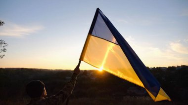 Erkek asker, güzel günbatımının arka planında Ukrayna bayrağıyla ayakta duruyor. Rus saldırısına karşı kazanılan zaferin şerefine mavi-sarı bayrak taşıyan Ukraynalı erkek asker..