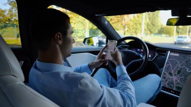 Başarılı bir iş adamı, otonom elektrikli araba kullanırken modern akıllı telefona göz atıyor. Şehir yolunda otomatik pilotla elektrikli araba sürerken telefon kullanan erkek girişimci..