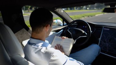 Erkek işadamı, şehir yolunda otomatik pilotla elektrikli araca binerken kitap okuyor. Başarılı bir iş adamı, kendi başına elektrikli araba sürerken bilgisini geliştiriyor..