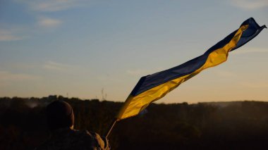 Askeri üniformalı genç adam arka planda güzel günbatımına karşı Ukrayna bayrağı sallıyor. Erkek Ukraynalı asker kırsal kesimde ulusal bayrağı kaldırdı. Rus saldırısına karşı zafer.