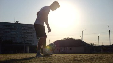 Günbatımında stadyumda futbol topu hokkabazlığı yapan profesyonel bir futbolcu. Genç adam yeşil sahada topa vuruyor. Sporcu arka planda güneş ışığıyla çayırda numaralar yapıyor. Serbest stil futbol kavramı.