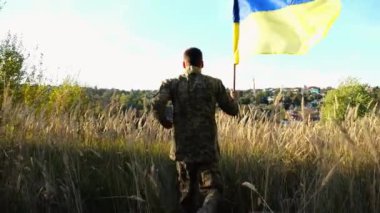 Ukrayna ordusunun erkek askeri açık havada mavi-sarı pankartla koşuyor. Kırsal kesimde Ukrayna bayrağı sallayan askeri üniformalı genç bir adam. Rus istila direnişi kavramı. Yavaş çekim