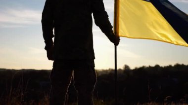 Ukraynalı asker gün batımında kırsal bölgede ulusal bayrağıyla ayakta duruyor. Rus saldırısına karşı zafer sembolü olarak Ukrayna bayrağı taşıyan askeri üniformalı bir erkek asker. İstila direnişi kavramı