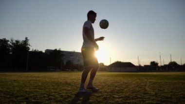 Günbatımında stadyumda futbol topu hokkabazlığı yapan profesyonel bir futbolcu. Genç adam yeşil sahada topa vuruyor. Sporcu arka planda güneş ışığıyla çayırda numaralar yapıyor. Serbest stil futbol kavramı.