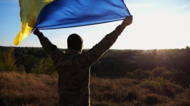 Üniformalı asker, arka planda güneş ışığına karşı Ukrayna bayrağıyla ayakta duruyor. Rus saldırısına karşı zafer anısına ulusal bayrağı kaldıran Ukraynalı erkek asker.