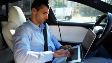 Şehir yolunda otonom bir otomatik araba sürerken dizüstü bilgisayar üzerinde çalışan yakışıklı bir girişimci. Erkek işadamı, otomatik pilotlu elektrikli aracın sürüşü sırasında dizüstü bilgisayarında yazı yazıyor..