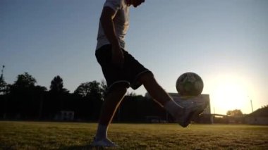 Günbatımında stadyumda futbol topu hokkabazlığı yapan profesyonel bir futbolcu. Genç adam yeşil sahada topa vuruyor. Sporcu arka planda güneş ışığıyla çayırda numaralar yapıyor. Serbest stil futbol kavramı.