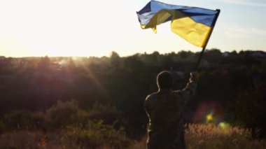Ukraynalı asker gün batımının arka planına ulusal bayrak sallıyor. Genç erkek asker kırsal kesimde bayrak kaldırdı. Rus saldırısına karşı zafer. Rusya ve Ukrayna arasındaki savaşın sonu..