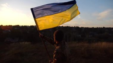 Ukrayna 'nın bayrağını gün batımında tepenin zirvesine sallayan Ukraynalı asker. Askeri üniformalı genç adam kırsal kesimde ulusal afişi kaldırdı. Rus saldırısına karşı zafer. Savaşın sonu..