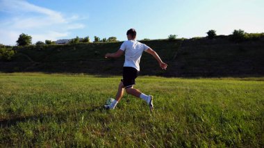 Üniformalı profesyonel futbolcu yeşil sahada koşarken topa numara yapıyor. Sporcu güneşli bir günde Meadow 'da koşarken topa vurur. Stadyumda antrenman yapan genç adam. Serbest stil futbol.