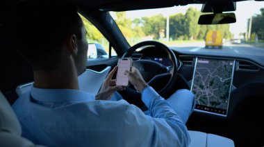 Başarılı bir iş adamı, otonom elektrikli araba kullanırken modern akıllı telefona göz atıyor. Şehir yolunda otomatik pilotla elektrikli araba sürerken telefon kullanan erkek girişimci..