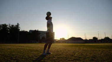 Günbatımında stadyumda futbol topu hokkabazlığı yapan profesyonel bir futbolcu. Genç adam yeşil sahada topa vuruyor. Sporcu arka planda güneş ışığıyla çayırda numaralar yapıyor. Serbest stil futbol kavramı.