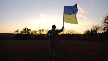 Alacakaranlıkta mavi-sarı bayrak taşıyan Ukrayna ordusunun askeri. Çayırda Ukrayna bayrağıyla koşan üniformalı genç bir erkek asker. Rus saldırganlığına karşı zafer.