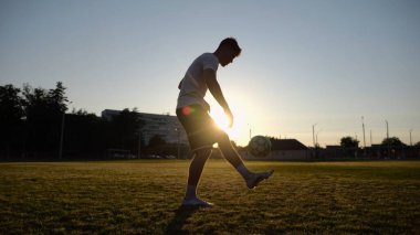 Günbatımında stadyumda futbol topu hokkabazlığı yapan profesyonel bir futbolcu. Genç adam yeşil sahada topa vuruyor. Sporcu arka planda güneş ışığıyla çayırda numaralar yapıyor. Serbest stil futbol kavramı.