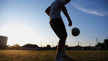 Günbatımında stadyumda futbol topu hokkabazlığı yapan profesyonel bir futbolcu. Genç adam yeşil sahada topa vuruyor. Sporcu arka planda güneş ışığıyla çayırda numaralar yapıyor. Serbest stil futbol kavramı.