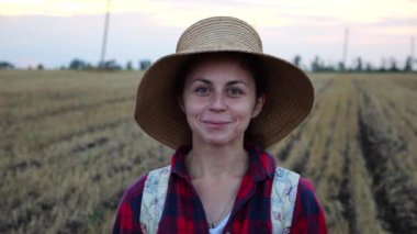 Mutlu gülümseyen dişi çiftçi buğday tarlasında kameraya bakıyor. Hasır şapkalı, arka planda arpa çayırı olan genç ve güzel tarımcı portresi. Tarım sektörü kavramı