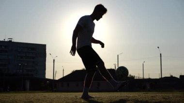 Günbatımında stadyumda futbol topu hokkabazlığı yapan profesyonel bir futbolcu. Genç adam yeşil sahada topa vuruyor. Sporcu arka planda güneş ışığıyla çayırda numaralar yapıyor. Serbest stil futbol kavramı.