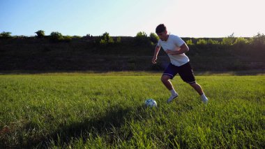 Üniformalı profesyonel futbolcu yeşil sahada koşarken topa numara yapıyor. Sporcu güneşli bir günde Meadow 'da koşarken topa vurur. Stadyumda antrenman yapan genç adam. Serbest stil futbol.