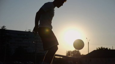 Günbatımında stadyumda futbol topu hokkabazlığı yapan profesyonel bir futbolcu. Genç adam yeşil sahada topa vuruyor. Sporcu arka planda güneş ışığıyla çayırda numaralar yapıyor. Serbest stil futbol kavramı.