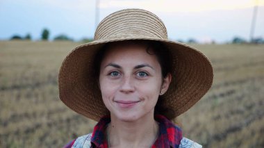 Mutlu gülümseyen dişi çiftçi buğday tarlasında kameraya bakıyor. Hasır şapkalı, arka planda arpa çayırı olan genç ve güzel tarımcı portresi. Tarım sektörü kavramı.