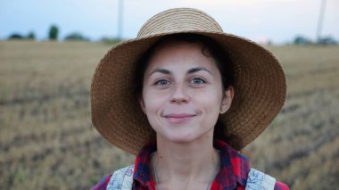 Mutlu gülümseyen dişi çiftçi buğday tarlasında kameraya bakıyor. Hasır şapkalı, arka planda arpa çayırı olan genç ve güzel tarımcı portresi. Tarım sektörü kavramı.