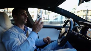 Şehir yolunda otomatik pilotla elektrikli otomobil sürerken akıllı telefondan konuşan erkek girişimci. Kendine güvenen işadamı, otomatik elektrikli araba kullanırken telefonla konuşuyor..