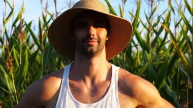 Mutlu gülümseyen erkek çiftçi mısır çayırının yanında kameraya bakar. Arkaplanda mısır tarlası olan hasır şapkalı genç ve yakışıklı tarımcı portresi. Tarım sektörü kavramı.