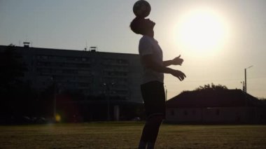 Sporcunun silueti gün batımında sahada topla numara yapıyor. Çayır 'da üniformalı profesyonel erkek futbolcu. Arka planda güneş ışığı var. Serbest stil futbol kavramı.